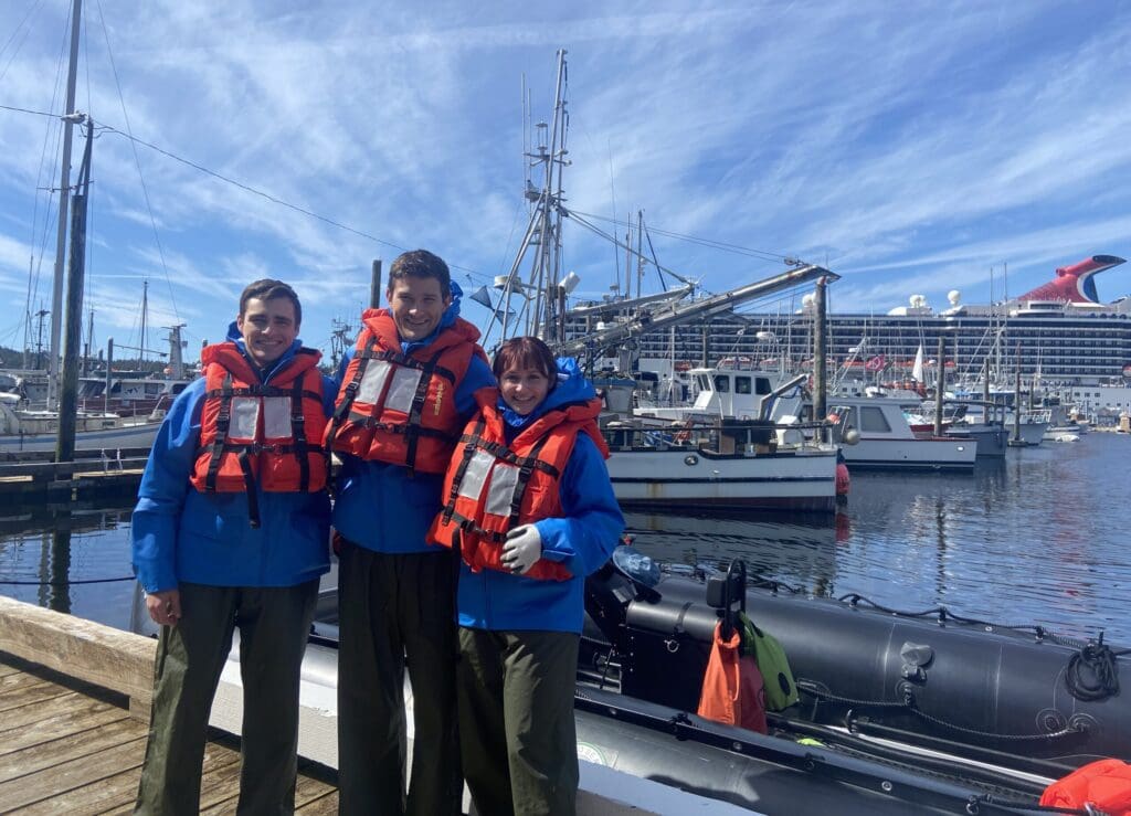 Three adults in rain gear and life jackets pose for a photo in front of a Zodiac in a harbor