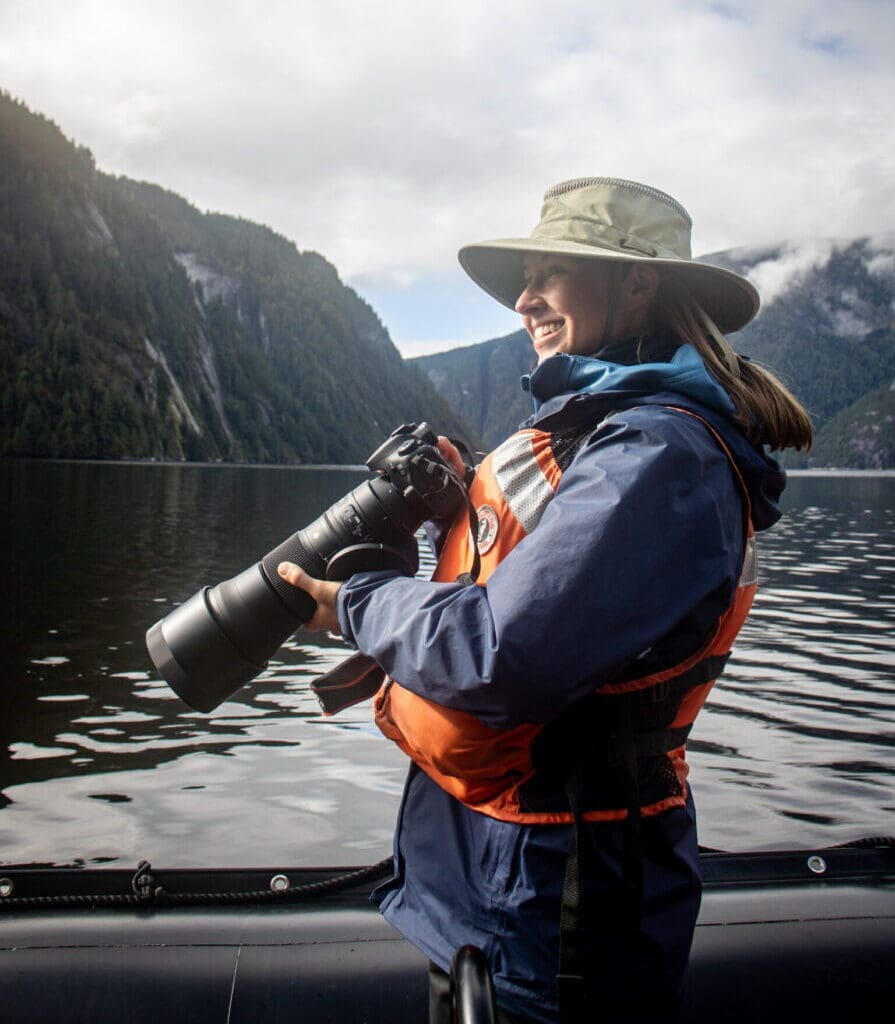 Woman in rain gear holding a camera standing in a Zodiac with a fjord in the background