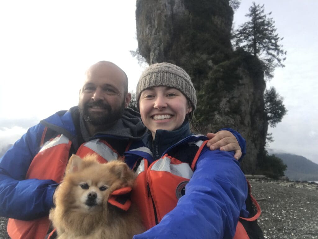 About Out to Sea Man, woman, and dog in life jackets pose in front of rock formation